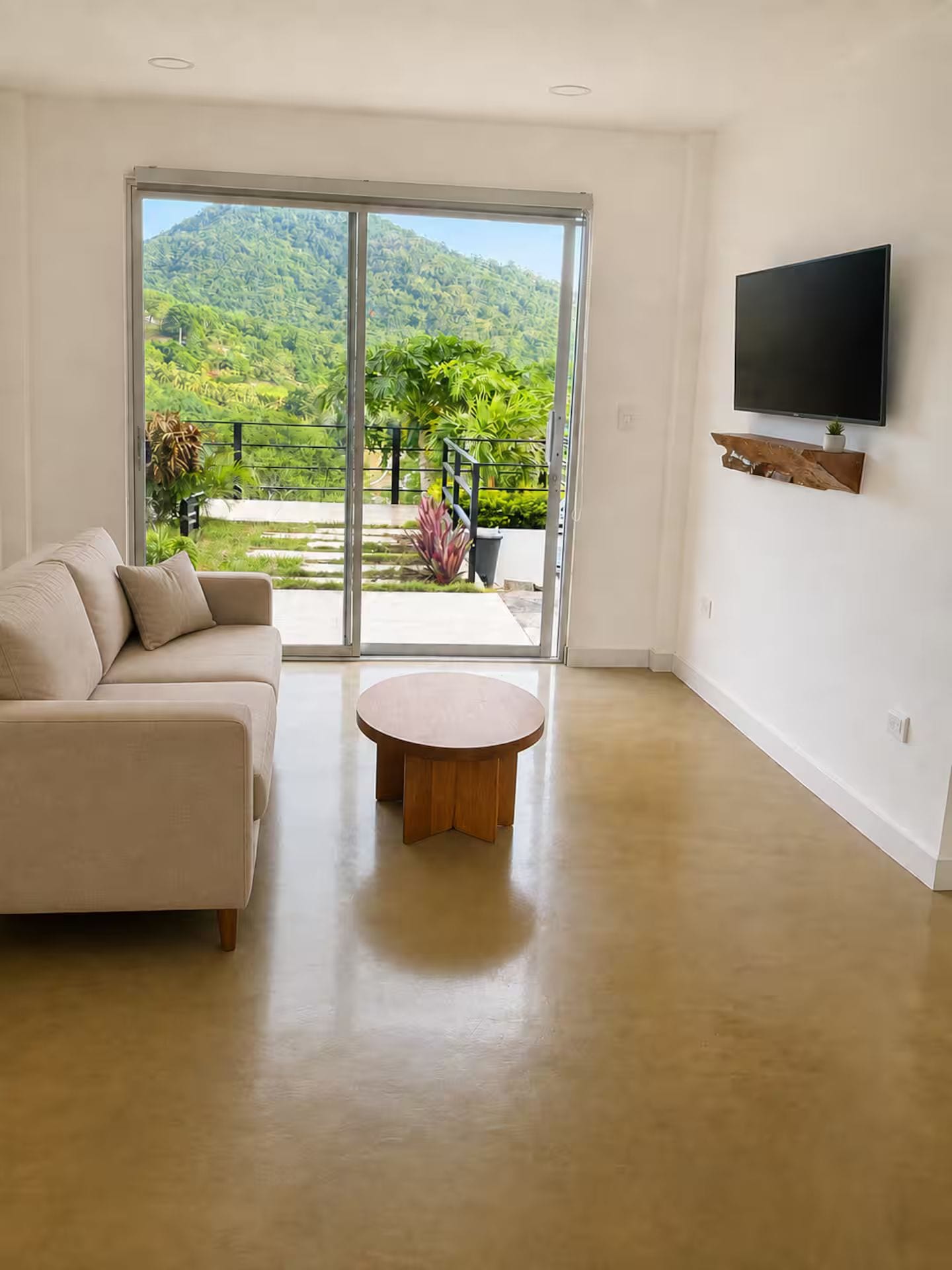 Una sala de estar moderna con un ventanal corredero que da a un balcón, un sofá color beige, una mesa redonda de madera y un televisor montado en la pared.