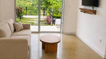 Una sala de estar moderna con un ventanal corredero que da a un balcón, un sofá color beige, una mesa redonda de madera y un televisor montado en la pared.