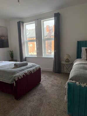 A bedroom with two beds, a window with curtains, a small table with a lamp, and a plant. - Comfortable 4BR House | Parking | Near City Ground (West Bridgford)