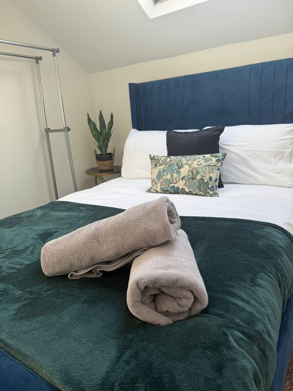 A neatly made bed with a green blanket, a blue headboard, and a potted plant on a bedside table.