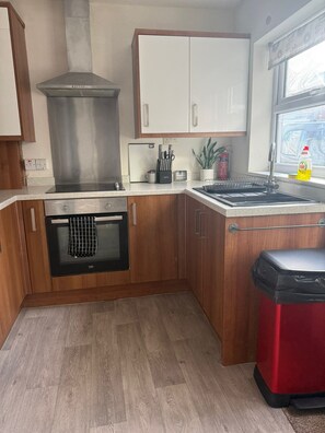 A compact kitchen with wooden cabinets, a stainless steel range hood, a built-in oven, and a window with a view. - Comfortable 4BR House | Parking | Near City Ground (West Bridgford)