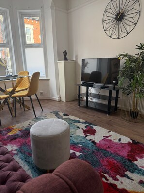 A living room with a television, a dining table with chairs, a colorful rug, and a potted plant. - Comfortable 4BR House | Parking | Near City Ground (West Bridgford)