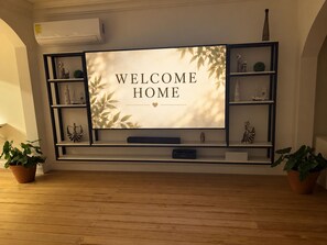 A flat-screen TV mounted on a wall with a 'WELCOME HOME' sign, flanked by shelves with decorative items and potted plants. - Horizonte Apartment: Nature and Pool (Sucio (San Juan del Sur))