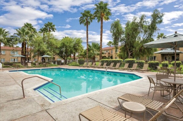 A swimming pool with lounge chairs and umbrellas, surrounded by palm trees and a building with balconies. - Palm Springs | Easy Access Studio Stay | Pool + Spa (Cathedral City)