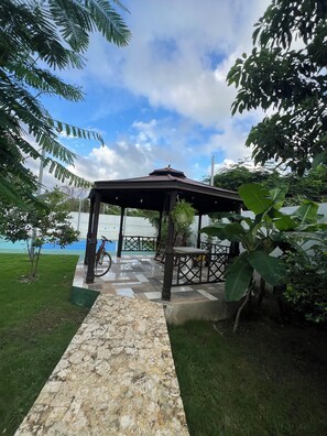 A gazebo with a tiled floor, surrounded by greenery and a clear sky.