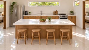 Een moderne keuken met een centraal eiland, houten krukken, een koelkast en een raam met uitzicht op groen.