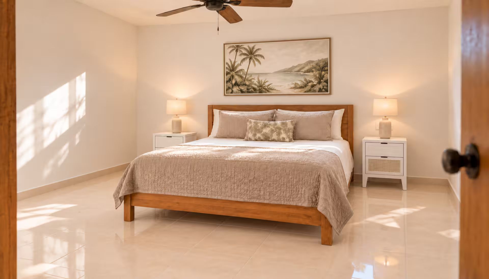 A bedroom with a wooden bed, two bedside tables with lamps, a ceiling fan, and a wall-mounted painting of a tropical scene.