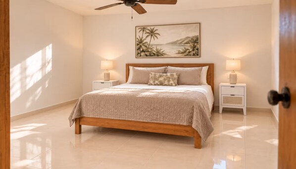 A bedroom with a wooden bed, two bedside tables with lamps, a ceiling fan, and a wall-mounted painting of a tropical scene.