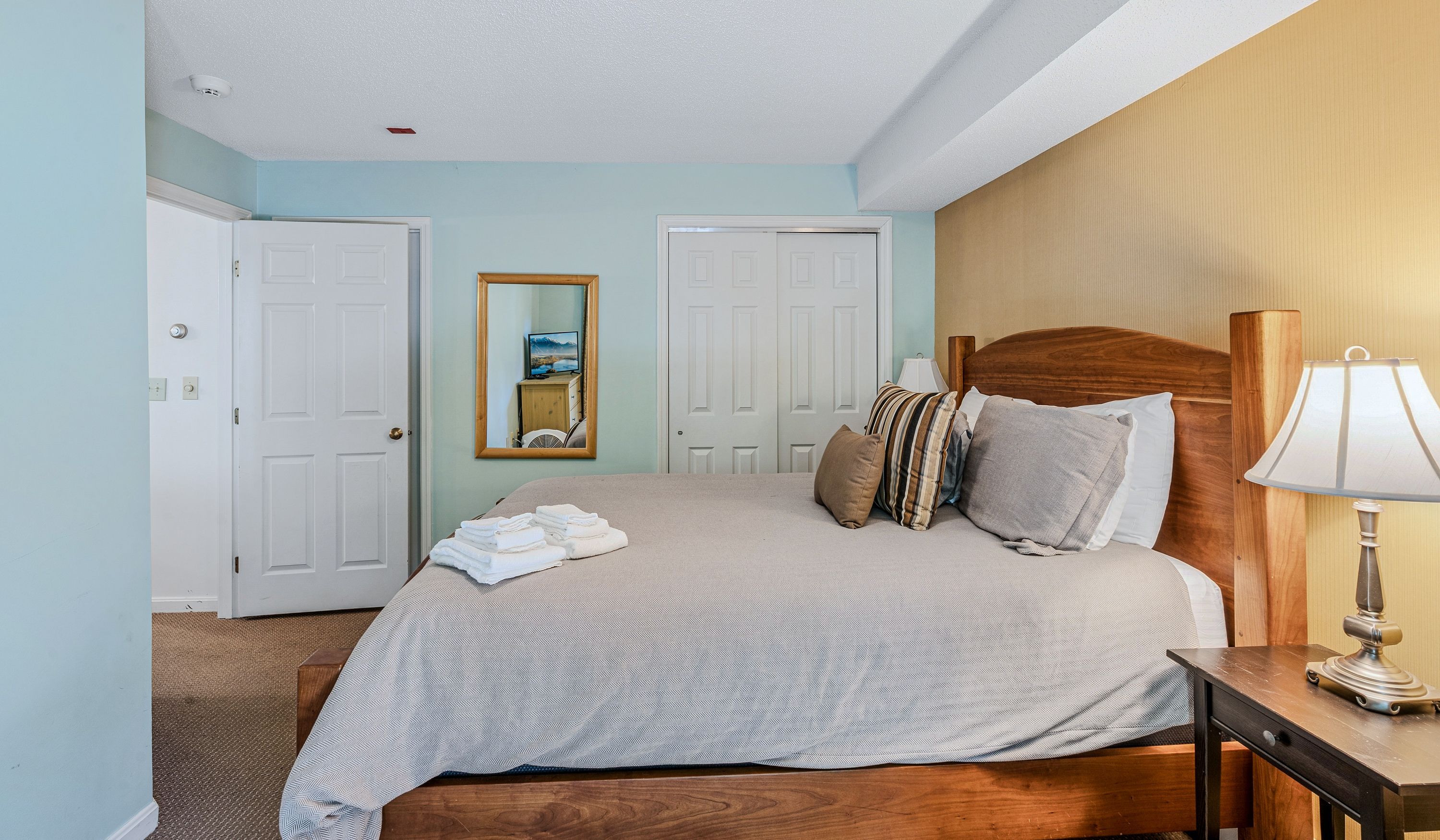 A hotel room with a bed, a bedside table with a lamp, a mirror, and two doors.