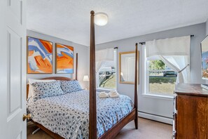 A four-poster bed with a floral bedspread, two abstract paintings, a bedside lamp, and a window with a view of a vehicle and greenery. - Okemo Ski Escape | Steps to the Slopes (Ludlow)
