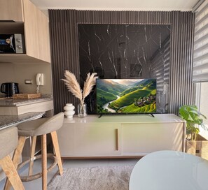 A modern living room with a flat-screen TV mounted on a wall, a dining area with a white table and chairs, and a kitchen area with a coffee machine and a phone. - Edificio Tempus by Domus Travels (San Miguel)