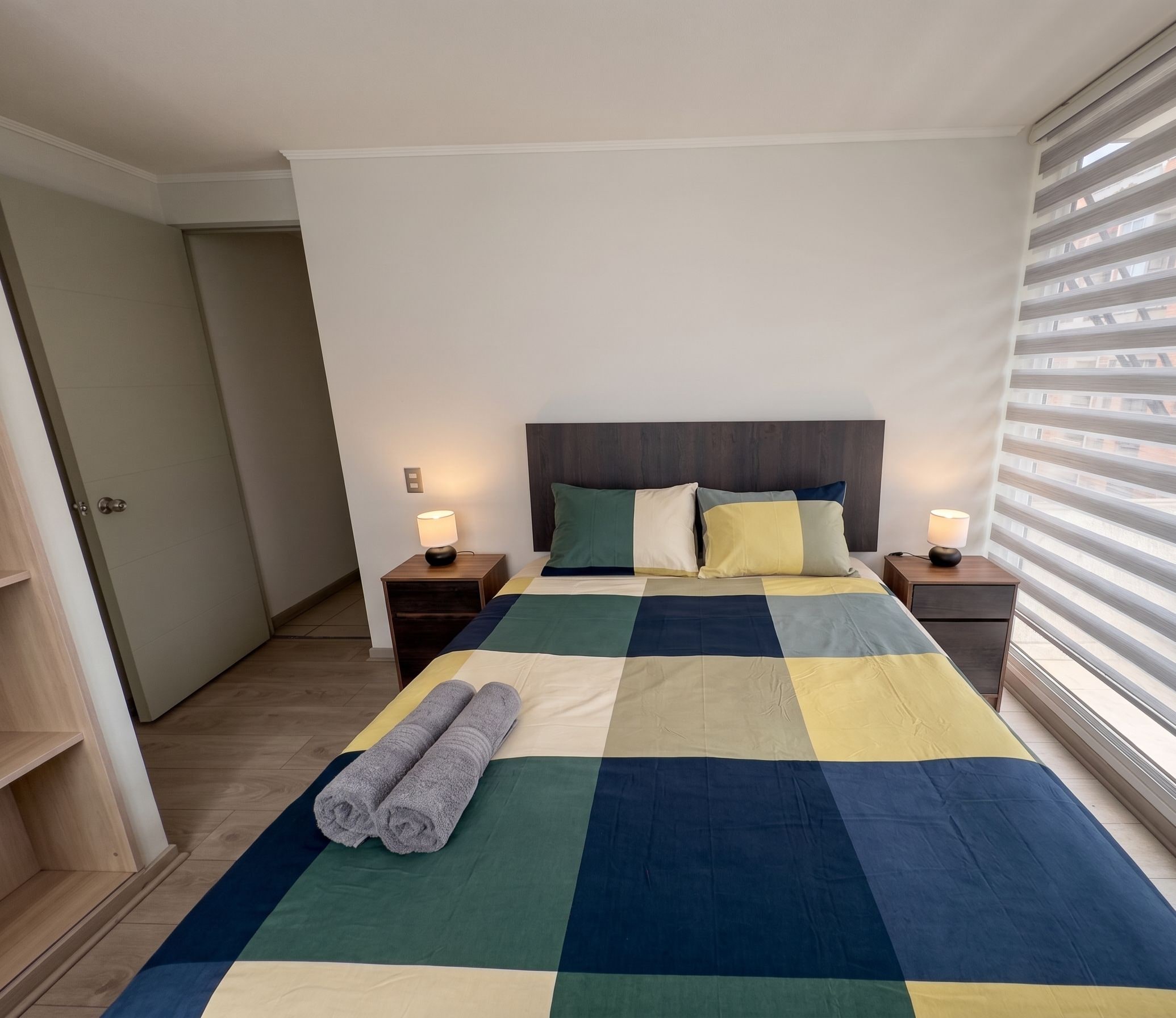 A bedroom with a checkered bedspread, two bedside lamps, a headboard, and a window with blinds.