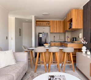 Een moderne keuken met houten kastjes, een inbouwoven en een koelkast. Er is een eethoek met een tafel en vier barkrukken.