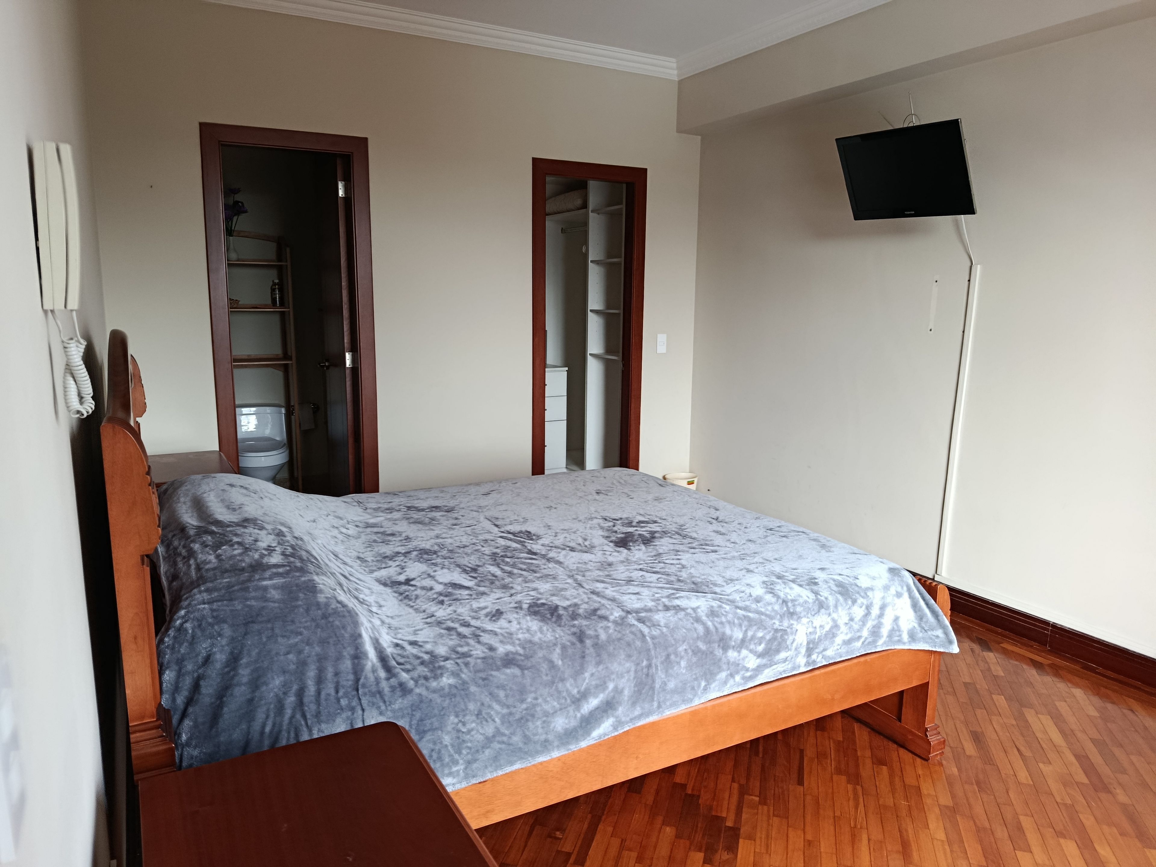 A bedroom with a bed, wooden floor, a TV mounted on the wall, and a wardrobe.