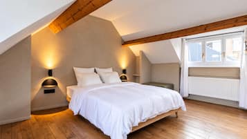 A bedroom with a sloped ceiling, a bed with white bedding, two wall-mounted light fixtures, and a window with curtains.