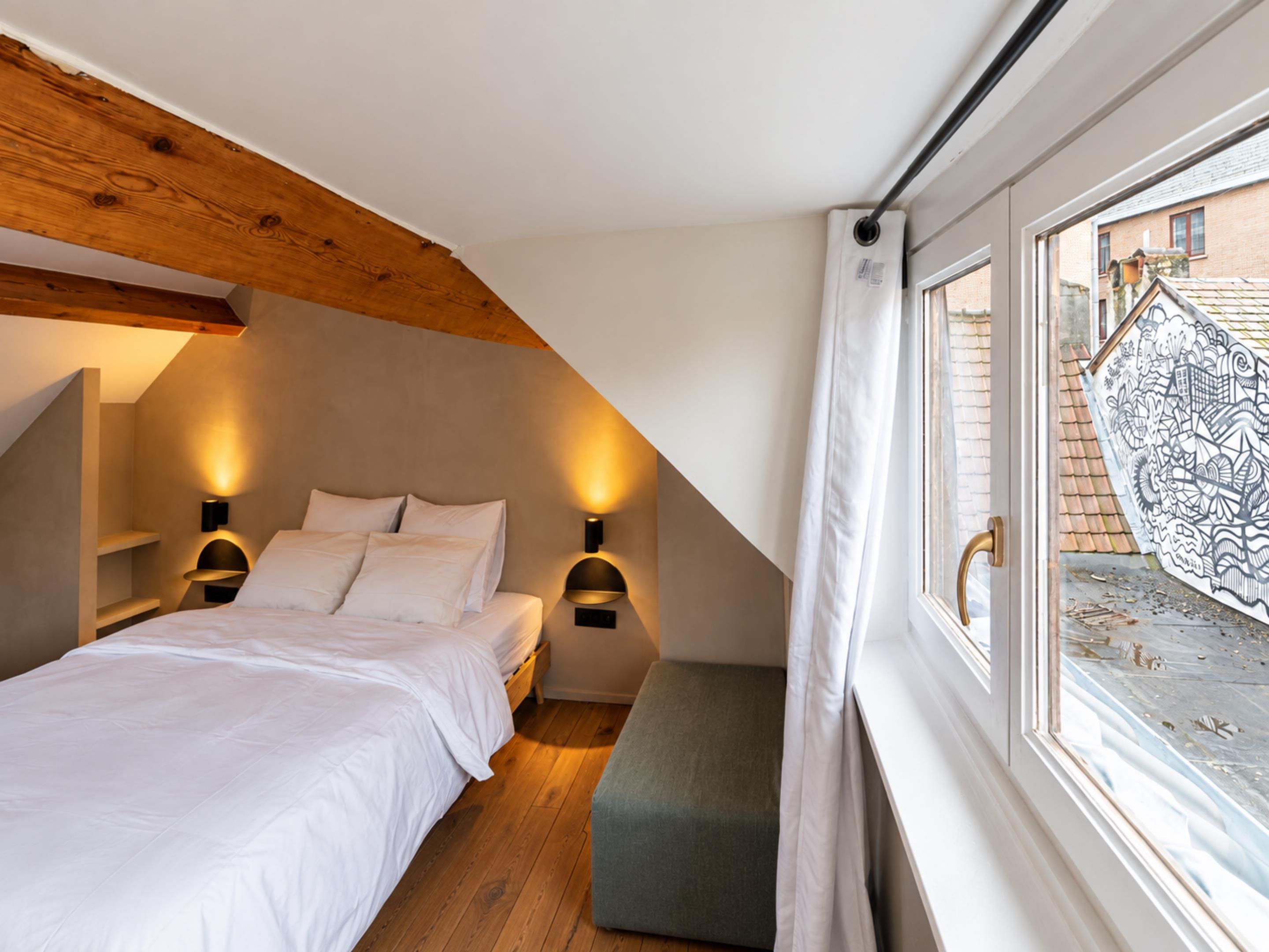 Ein Schlafzimmer mit schrägen Wänden, einem Bett mit weißer Bettwäsche, zwei Wandleuchten und einem Fenster mit Blick auf ein Gebäude.