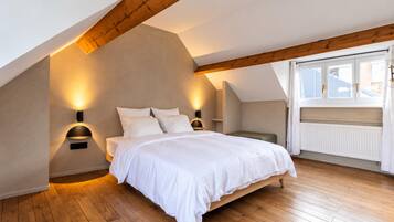 Ein Schlafzimmer mit schrägem Dach, Holzbalken, einem Bett mit weißer Bettwäsche, zwei Wandleuchten und einem Fenster mit Vorhängen.
