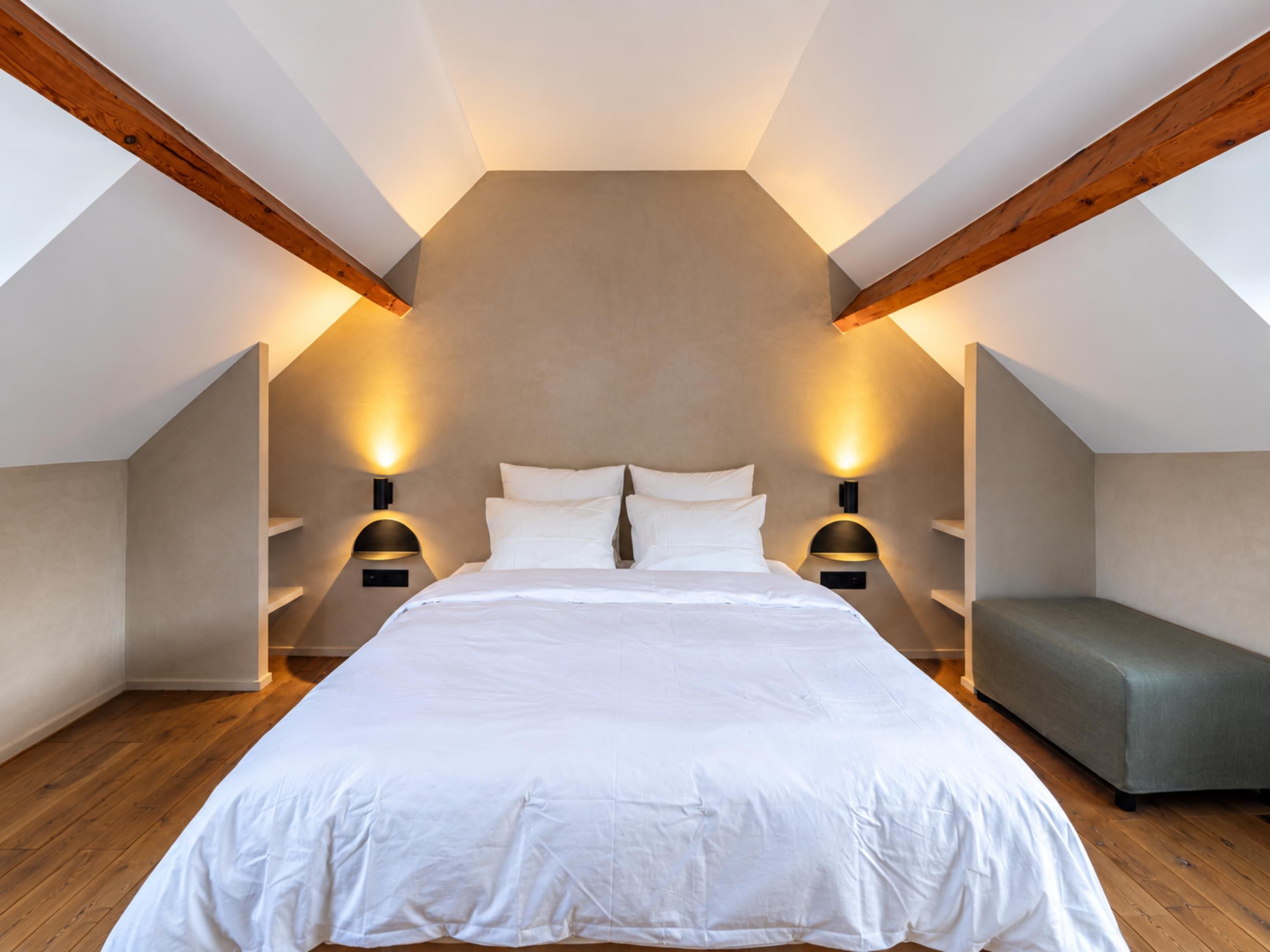 A bedroom with a large bed, two bedside tables with lamps, a bench, and exposed wooden beams.