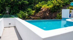 Une piscine dotée d’une terrasse blanche, entourée d’une végétation luxuriante et bordée d’une falaise rocheuse.