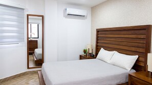 Une chambre d’hôtel comprenant un lit, une table de chevet avec une lampe, une petite table avec une horloge, un miroir et un appareil de climatisation.