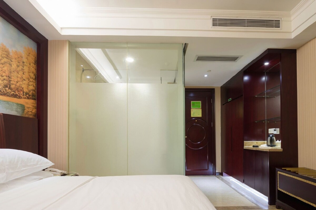 Ein Hotelzimmer mit einem Bett, einer Glastür zum Badezimmer, einem Schminktisch mit Spiegel und einem Bild an der Wand.