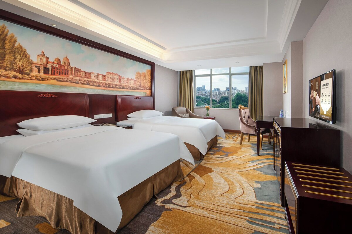 Ein Hotelzimmer mit einem großen Bett, einem Schreibtisch, einem Sessel, einem Fernseher und einem Gemälde an der Wand.