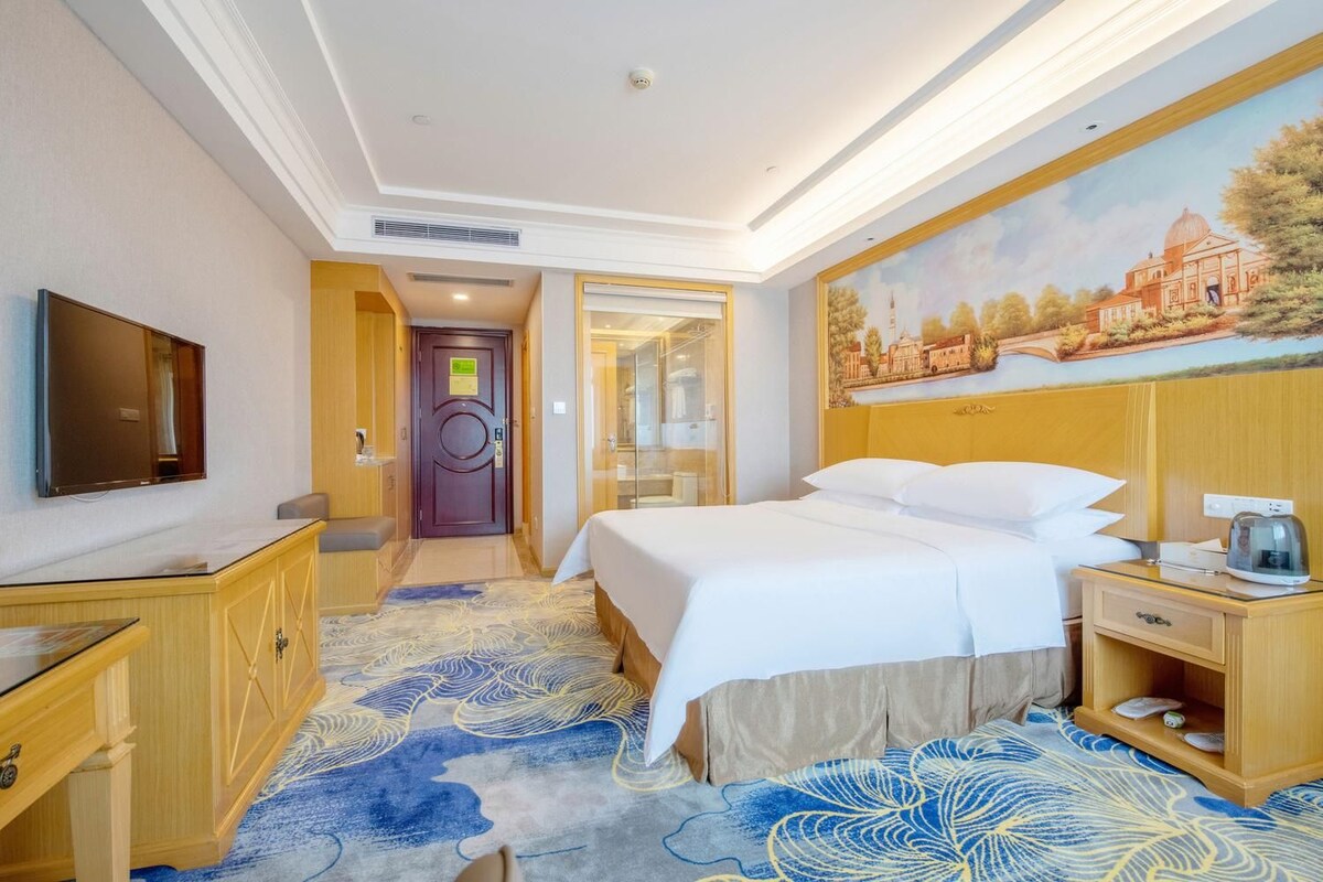 A hotel room with a bed, a desk, a television, and a painting on the wall.