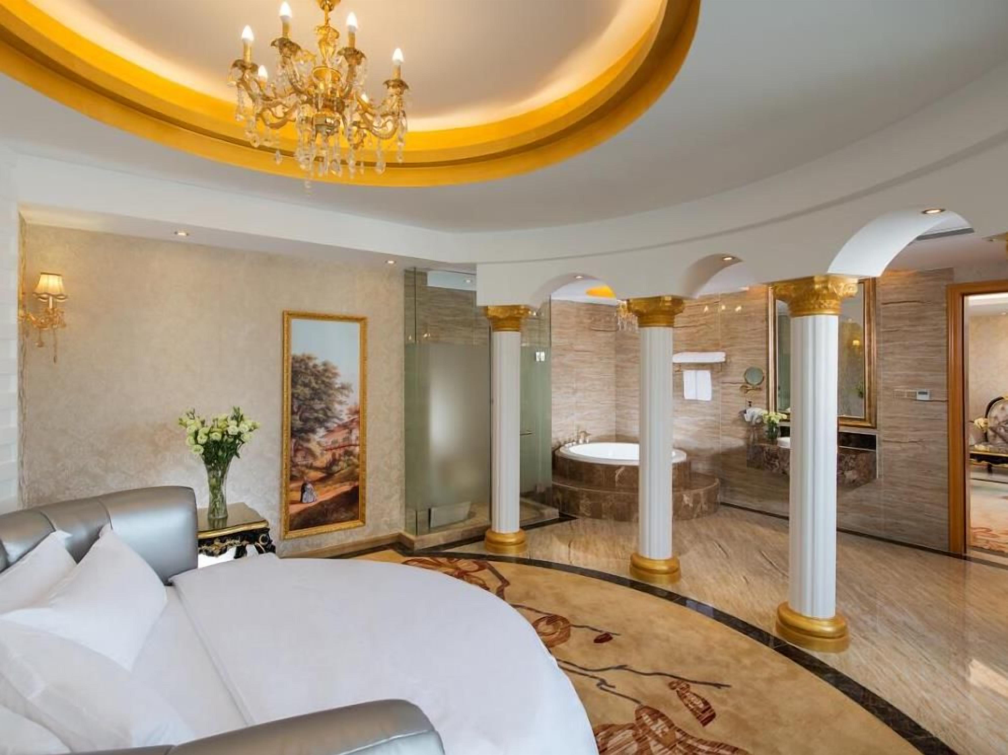 A luxurious hotel room with a large bed, a chandelier, a painting, a bathroom with a bathtub, and a marble floor.