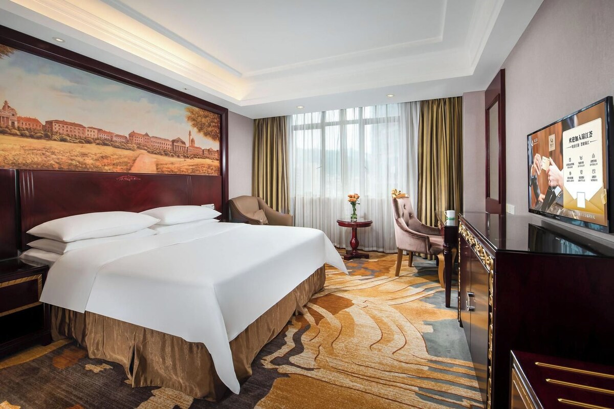 Ein Hotelzimmer mit einem großen Bett, einem Schreibtisch, einem Stuhl, einem Fernseher und einem Gemälde an der Wand.