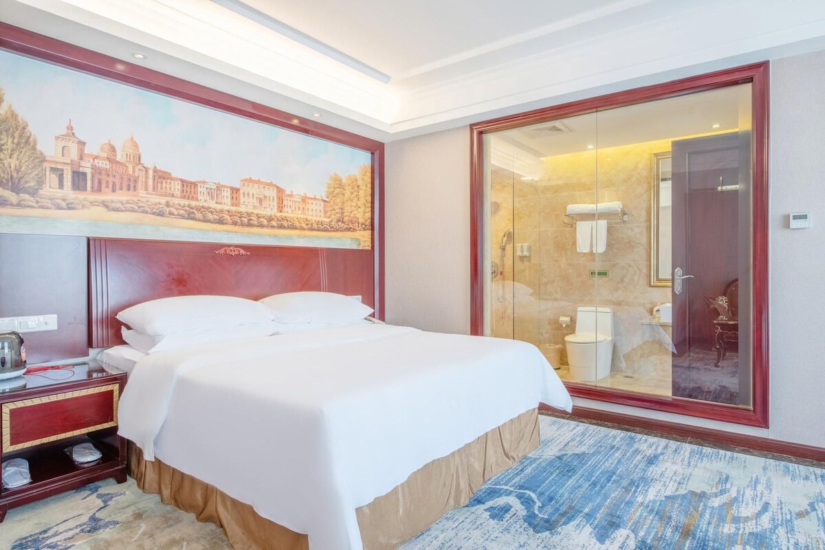 Ein Hotelzimmer mit einem großen Bett, einem Nachttisch, einem Stadtbild und einem durch Glas geschützten Badezimmer sowie einem spiegel mit rotem Rahmen.