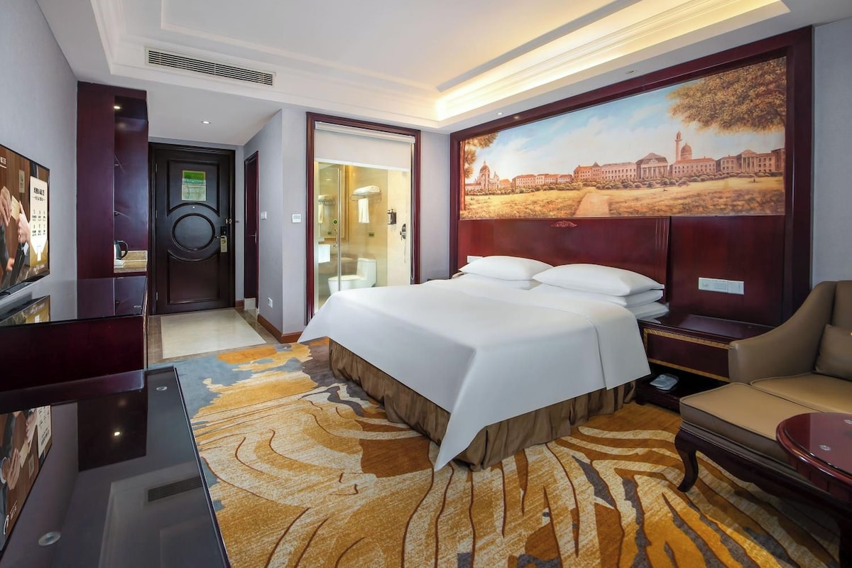 A hotel room with a bed, a seating area, a shower, and a large painting on the wall.