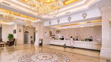 A hotel lobby with a reception desk, chandeliers, a decorative floor pattern, and a clock on the wall.