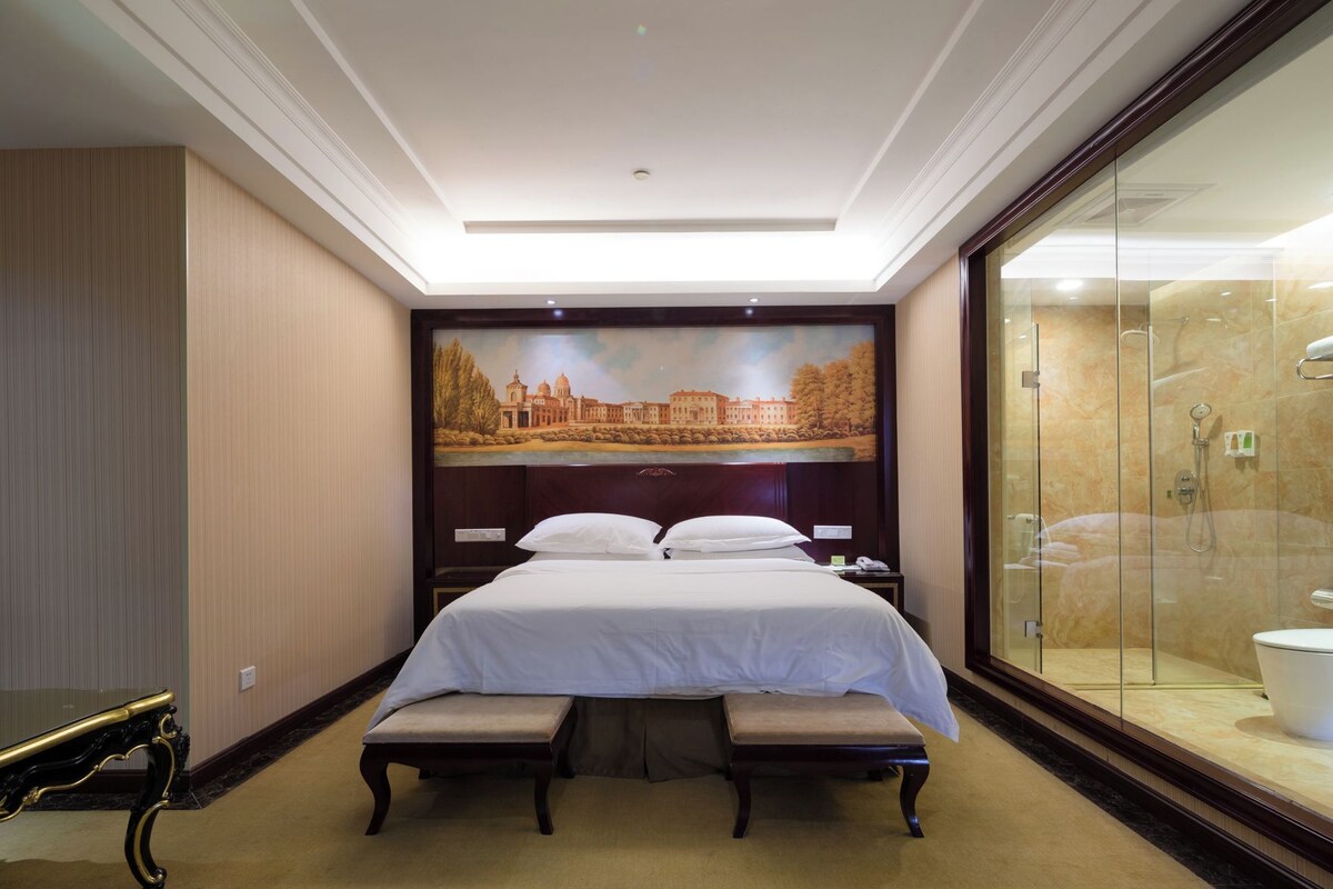 Een hotelkamer met een groot bed, een bankje, een douchecabine van glas en een muurschildering.