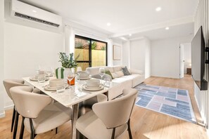 A modern dining area with a table set for four, a sofa, and a flat-screen TV mounted on the wall. - Brand-New Home | Walk to Beach & Dining | full A/C (Surfers Paradise)
