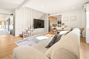 A modern living room with a sofa, a TV, and a dining area with chairs and a table. - Brand-New Home | Walk to Beach & Dining | full A/C (Surfers Paradise)