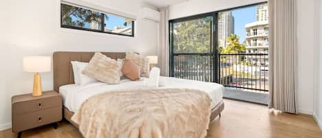 Quarto com cama grande, mesa de cabeceira, janela com vista para edifícios e varanda com guarda-corpo.