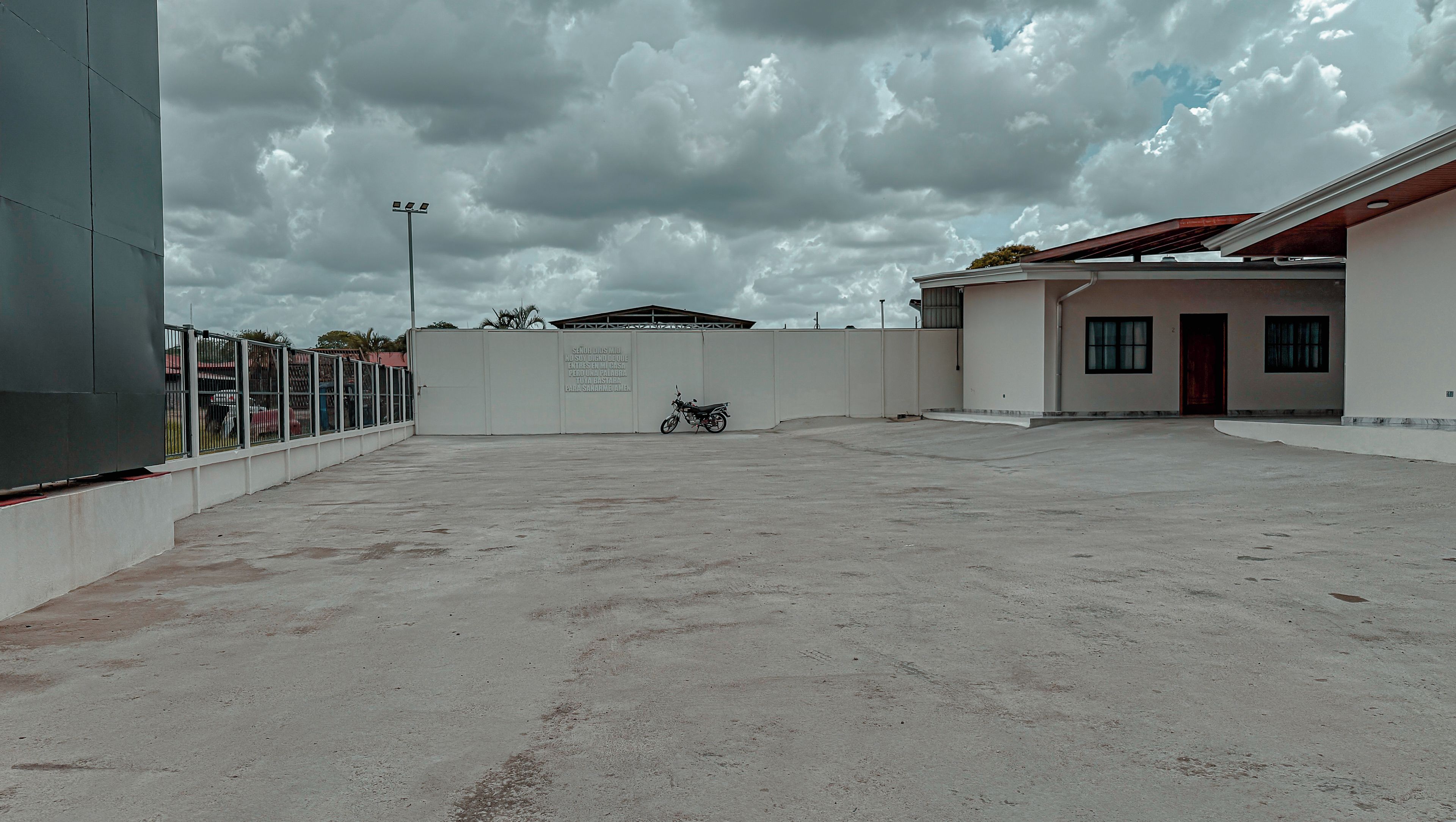 An empty parking lot with a motorcycle, a fenced area, and a building with a flat roof.