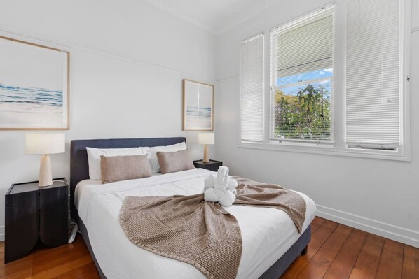 A bedroom with a bed, two bedside tables, a window with blinds, and two framed pictures on the wall. - Character Retreat| Prime location|Pool & Game Room (Ascot)