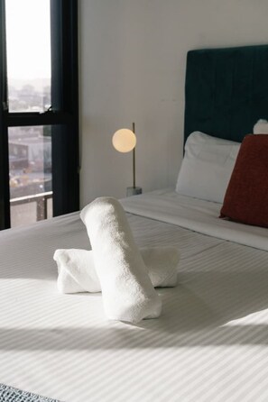 Ein ordentlich bezogenes Bett mit weißen Leinentüchern, einer Nachttischlampe und Blick durch ein Fenster.