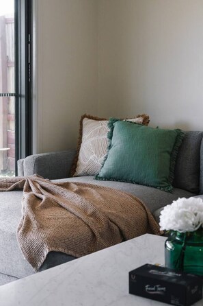 Un salon avec un canapé gris, un plaid à motifs et un vase vert contenant des fleurs blanches, posé sur une table.