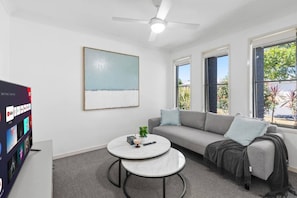 Ein modernes Wohnzimmer mit einem grauen Sofa, zwei runden Tischen, einem Deckenventilator und einem großen Fenster mit Blick ins Grüne.