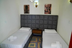 Una habitación con dos camas individuales, un cabecero acolchado gris, una mesita de noche de madera pequeña y un suelo con un estampado colorido.