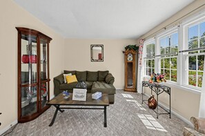 A living room with a sofa, a coffee table, a cabinet, a clock, and a window with a view of greenery. - Near MCAS New River - Peaceful Home w Patio (Jacksonville)