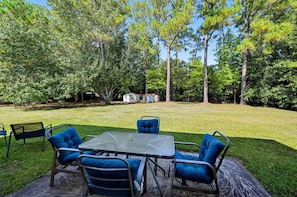 Une terrasse extérieure avec une table en métal et des chaises rembourrées bleues, entourée d’une pelouse verdoyante et d’arbres.