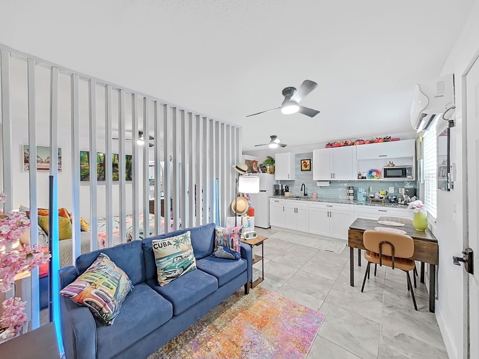 A modern living room with a blue sofa, a dining area with a wooden table and chairs, and a kitchen with white cabinets and a microwave.