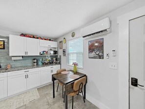 Cozinha compacta com armários brancos, uma mesa de jantar com cadeiras e uma janela com persianas.