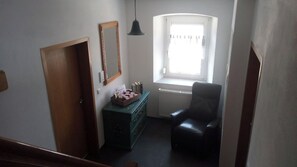 Habitació amb una porta de fusta, un armari amb calaixos, una cadira negra, una finestra amb cortines i una làmpada penjant.