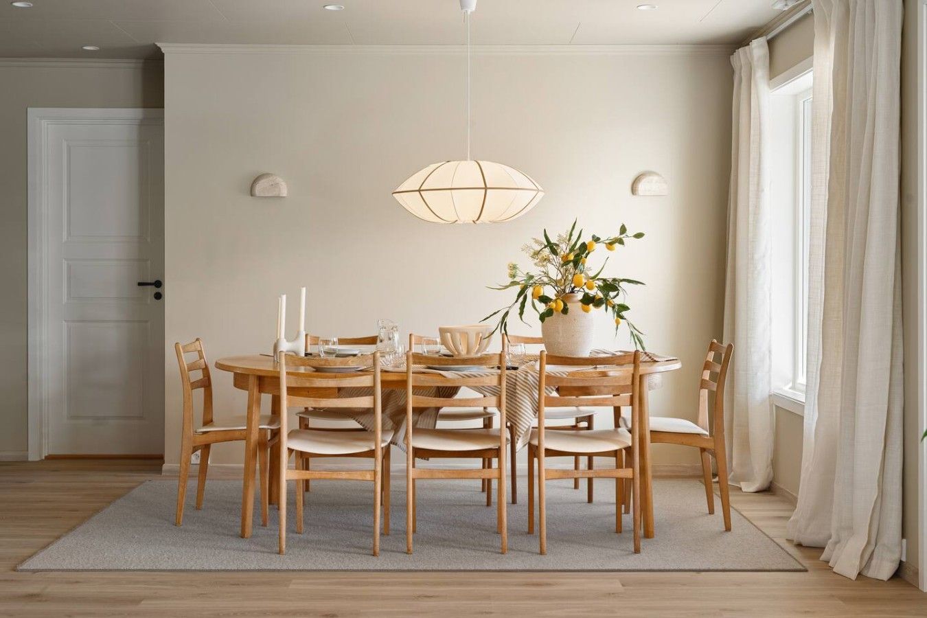 A dining area with a round table, six chairs, a hanging light fixture, and a potted plant with yellow flowers.