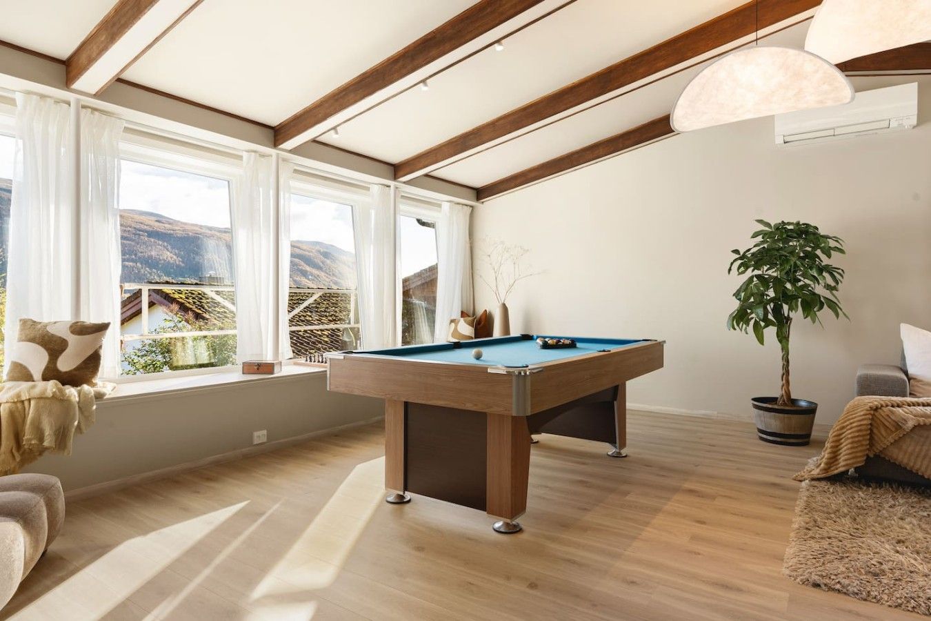 A room with a pool table, a sofa, a potted plant, and large windows with a view of a mountainous landscape.