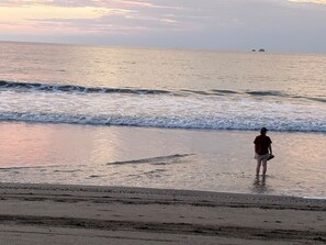 A beach scene with a person standing in the water, waves approaching the shore, and a distant landmass. - Tropical Villa • Private Pool • Near Conchal & Flamingo (Brasilito)